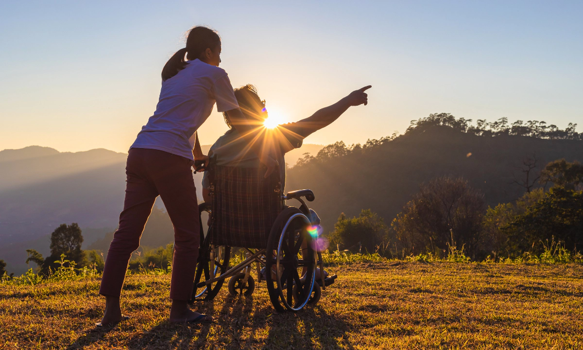 A woman with a ponytail and a man who is a wheelchair user in front of a glowing sunrise and background of mountains and trees. The man is pointing to something out of frame, and the image conveys travel and adventure. A woman with a ponytail and a man who is a wheelchair user in front of a glowing sunrise and background of mountains and trees. The man is pointing to something out of frame, and the image conveys travel and adventure.