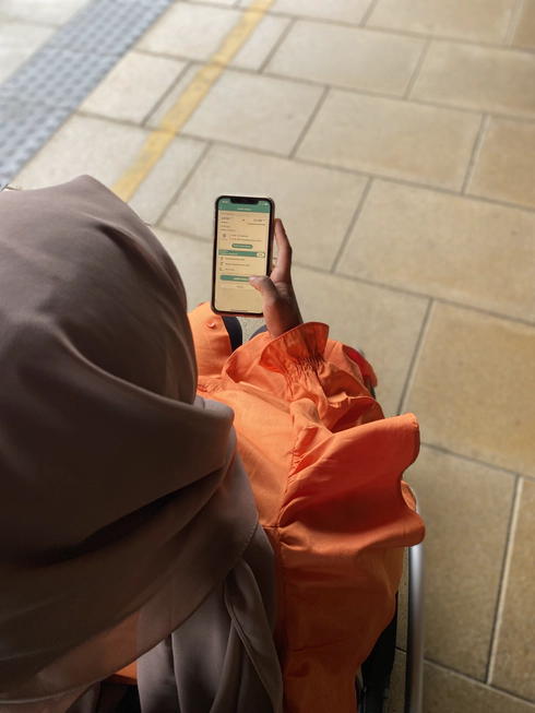 woman in a brown headscarf and orange long sleeved dress holding a smartphone. The woman is a wheelchair user and she is on a train platform. Her phone screen shows the Passenger Assistance app.