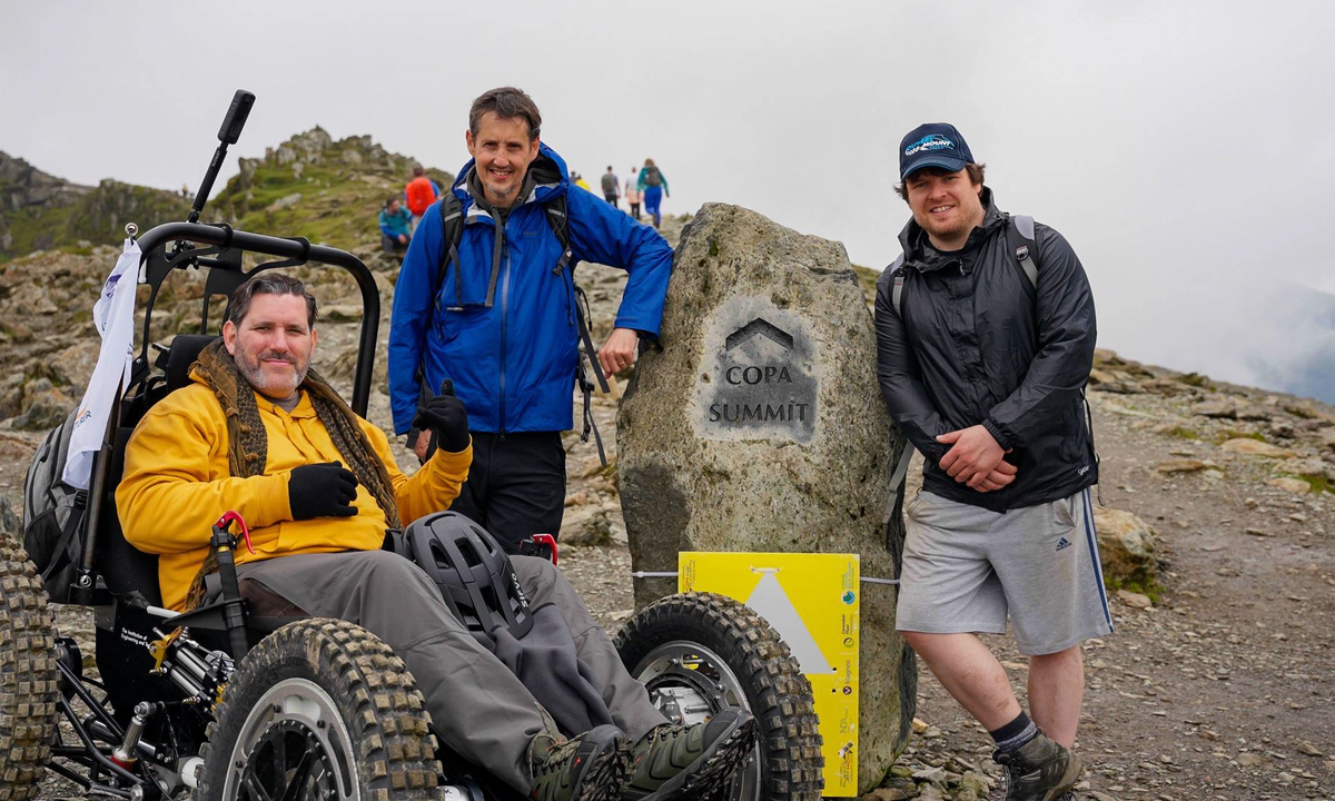 Nick Wilson in RockClimber beside the Copa Summit sign on Mount Snowdon. Nick is smiling, wearing a yellow hoodie, grey trousers, and black gloves. Nick Wilson in RockClimber beside the Copa Summit sign on Mount Snowdon. Nick is smiling, wearing a yellow hoodie, grey trousers, and black gloves.