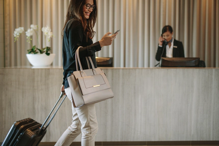 A woman smiling at her smart phone with a suitcase and handbag in front of a hotel check-in desk.
