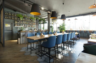 A photo of a restaurant with a long table, blue chairs, ceiling lamps, and wine bar.