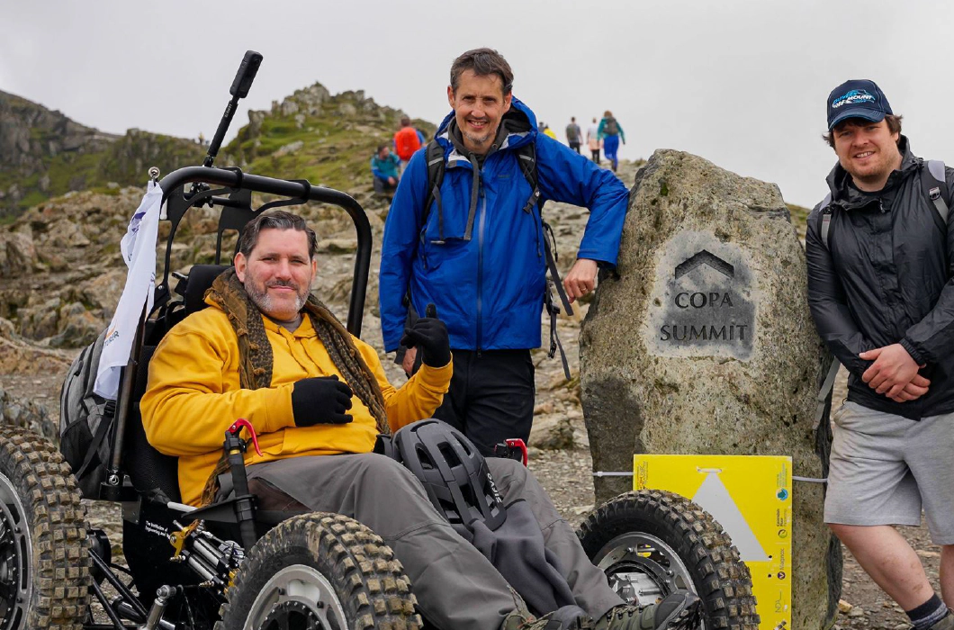 Nick Wilson in RockClimber beside the Copa Summit sign on Mount Snowdon. Nick is smiling, wearing a yellow hoodie, grey trousers, and black gloves.