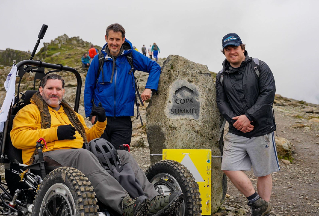 Nick Wilson in RockClimber beside the Copa Summit sign on Mount Snowdon. Nick is smiling, wearing a yellow hoodie, grey trousers, and black gloves.