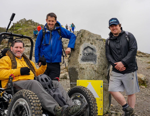 Nick Wilson in RockClimber beside the Copa Summit sign on Mount Snowdon. Nick is smiling, wearing a yellow hoodie, grey trousers, and black gloves.