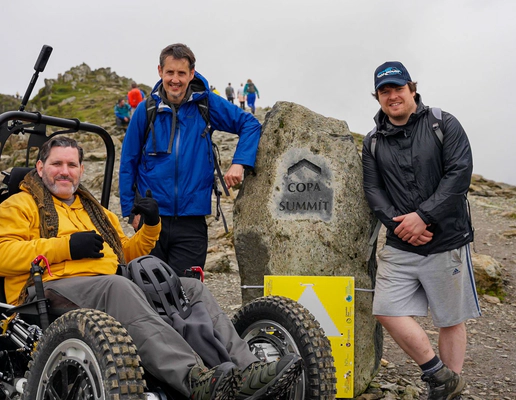 Nick Wilson in RockClimber beside the Copa Summit sign on Mount Snowdon. Nick is smiling, wearing a yellow hoodie, grey trousers, and black gloves. Nick Wilson in RockClimber beside the Copa Summit sign on Mount Snowdon. Nick is smiling, wearing a yellow hoodie, grey trousers, and black gloves.