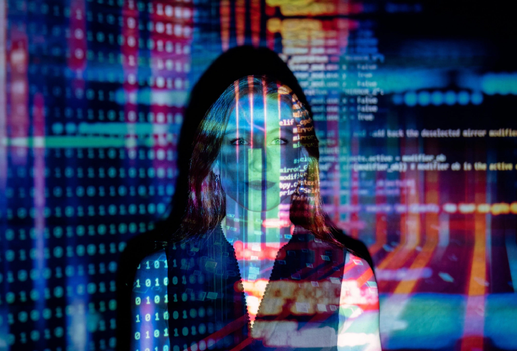 Photo of a woman standing in front of a wall facing the camera with colourful lights, lines of code, text and shapes projected onto her to convey tech.