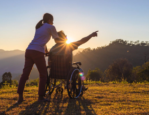 A woman with a ponytail and a man who is a wheelchair user in front of a glowing sunrise and background of mountains and trees. The man is pointing to something out of frame, and the image conveys travel and adventure.