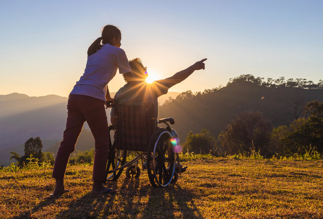 A woman with a ponytail and a man who is a wheelchair user in front of a glowing sunrise and background of mountains and trees. The man is pointing to something out of frame, and the image conveys travel and adventure.