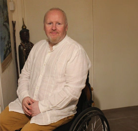 Jamie, a white man with short hair and facial hair, wearing a white shirt and tan trousers, sitting in his wheelchair, smiling at the camera.