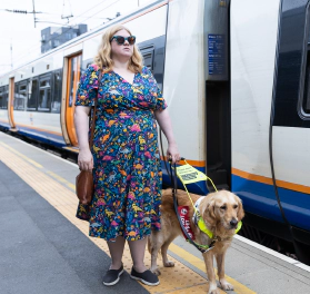 Dr Amy Kavanagh, a white blonde woman wearing a colourful dress and black glasses, standing on tactile paving beside her guide dog on a train station platform, with a train to their side
