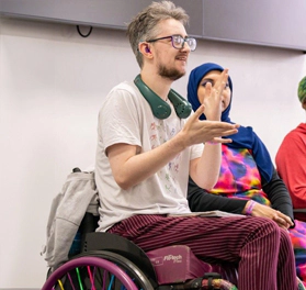 Jasper Armitage-Williams, a white man with short brown hair and facial hair, wearing black glasses, a white t-shirt and red trousers, sitting in his wheelchair, and signing towards someone out of view