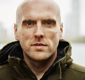 Simon Wheatcroft, a white man with a shaved head wearing a green coat and looking into the camera