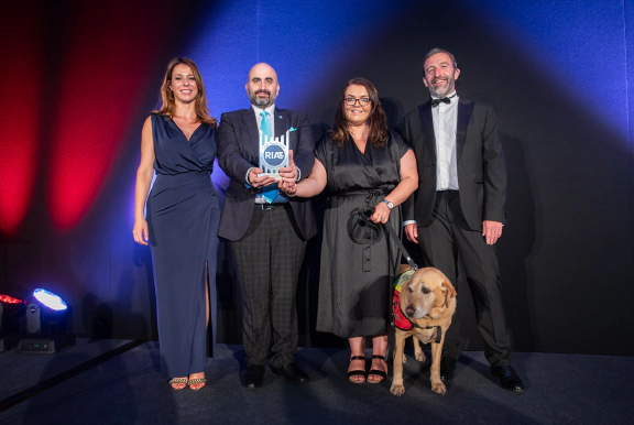 Transreport's Commercial Manager Joseph Mastracci, and Transreport's Accessibility and Inclusion Manager Emma Partlow holding the RIA RISE Award. Emma holds her assistance dog Luna on a black lead. Everyone in the photo is smiling proudly. 