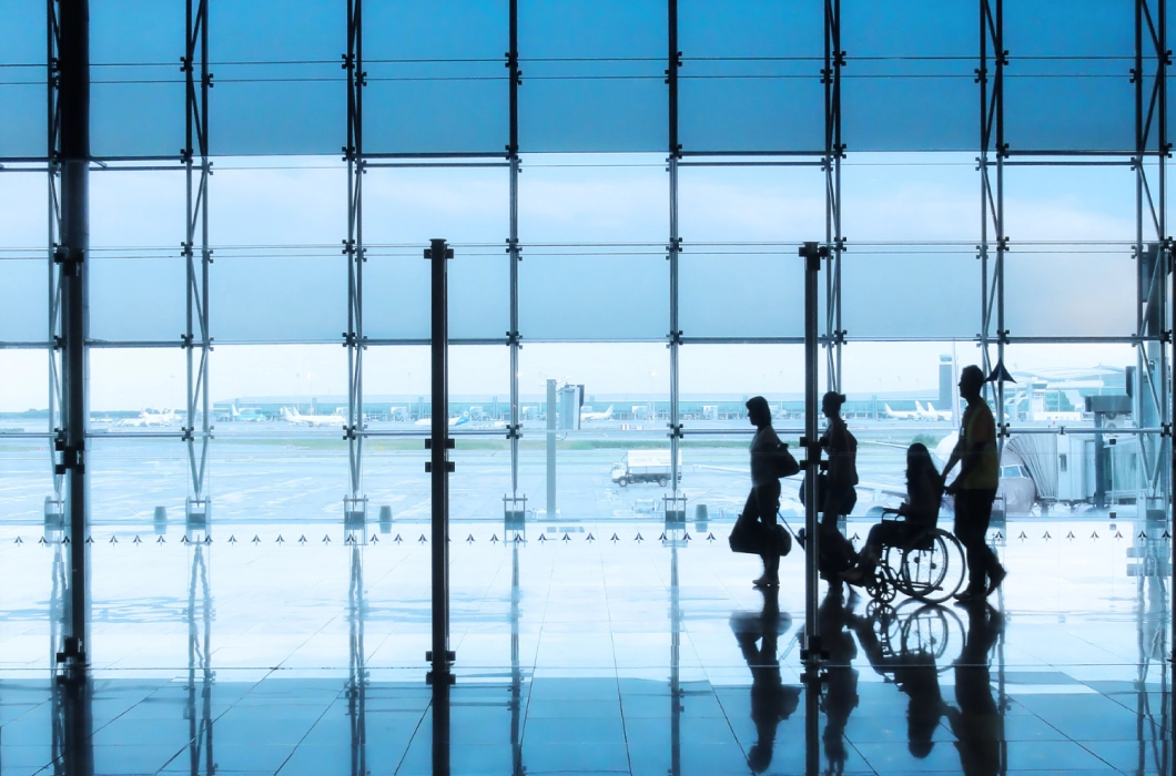Silhouettes of four people, three walking and one in a wheelchair, in an airport lounge with an aeroplane runway in the background.