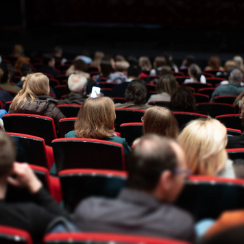 A photo of people at a venue seated in red chairs in the audience. A photo of people at a venue seated in red chairs in the audience.