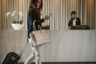 A woman smiling at her smart phone with a suitcase and handbag in front of a hotel check-in desk.