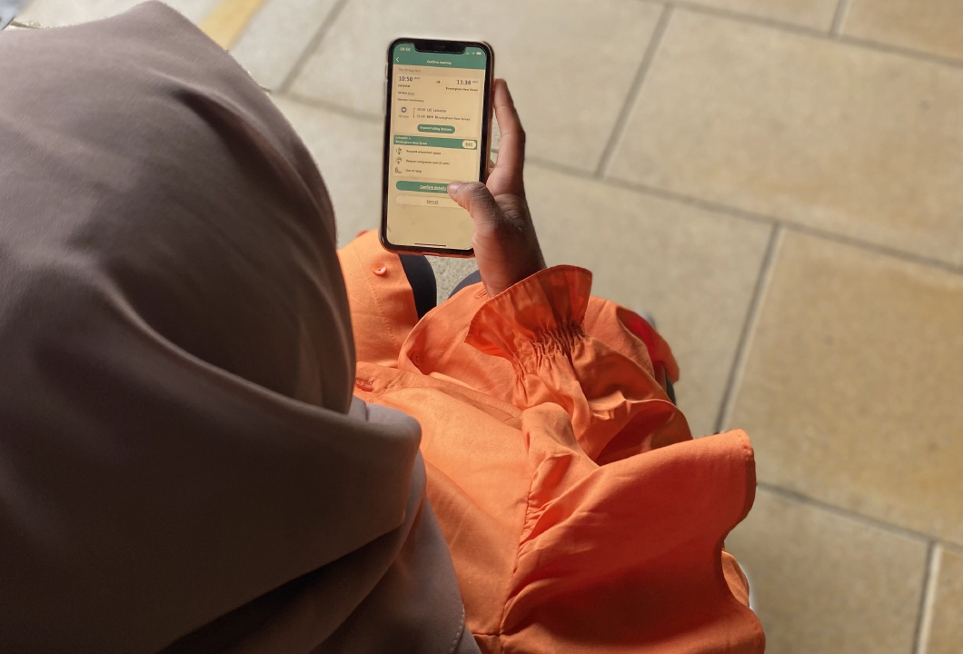 woman in a brown headscarf and orange long sleeved dress holding a smartphone. The woman is a wheelchair user and she is on a train platform. Her phone screen shows the Passenger Assistance app.