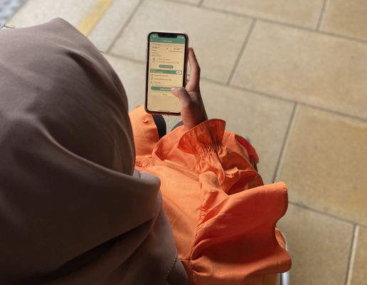 woman in a brown headscarf and orange long sleeved dress holding a smartphone. The woman is a wheelchair user and she is on a train platform. Her phone screen shows the Passenger Assistance app.