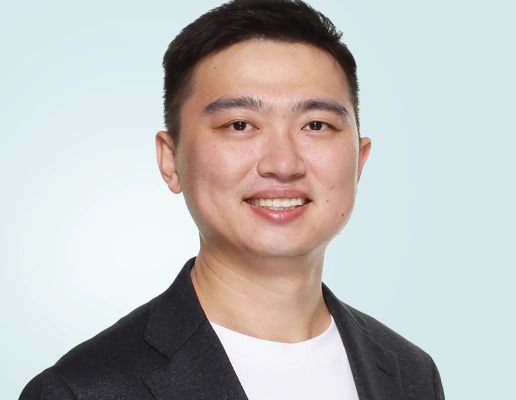 جاي شين، رجل آسيوي ذو شعر أسود قصير، والرئيس التنفيذي لشركة Transreport، يرتدي بدلة رمادية ويقف على منصة التحدث .