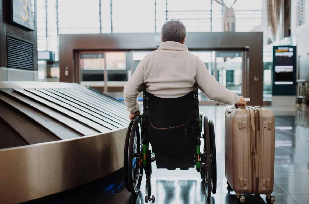 A man wearing a white jumper sitting in a wheelchair with his back to the camera, holding a suitcase on wheels in his right hand, in an airport.