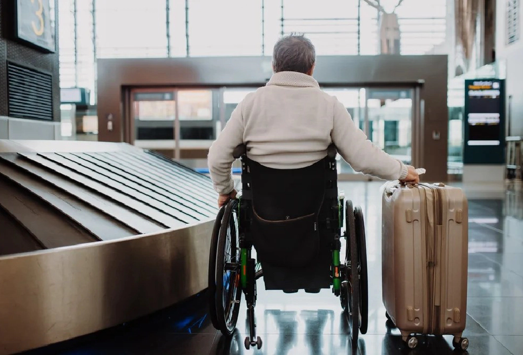 A man wearing a white jumper sitting in a wheelchair with his back to the camera, holding a suitcase on wheels in his right hand, in an airport.