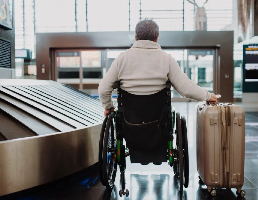 A man wearing a white jumper sitting in a wheelchair with his back to the camera, holding a suitcase on wheels in his right hand, in an airport.