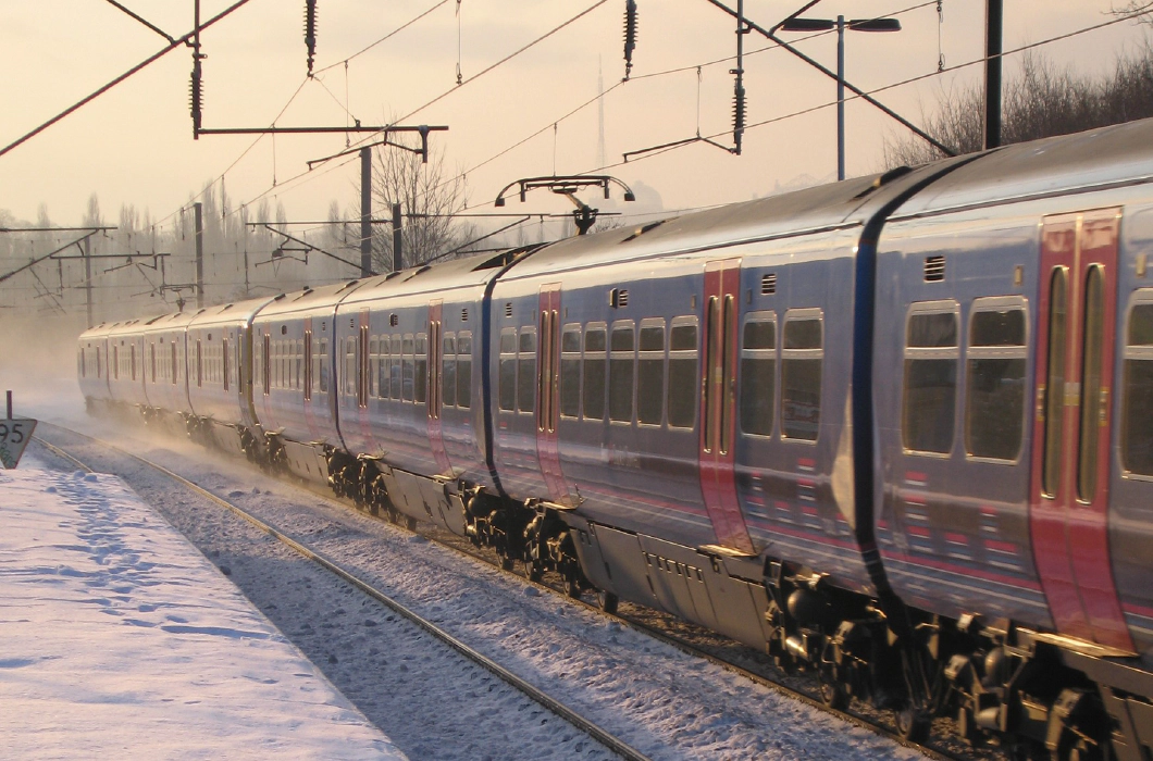 Photo of a blue and red train on a snowy track beside a snowy platform with an orange sky behind it.