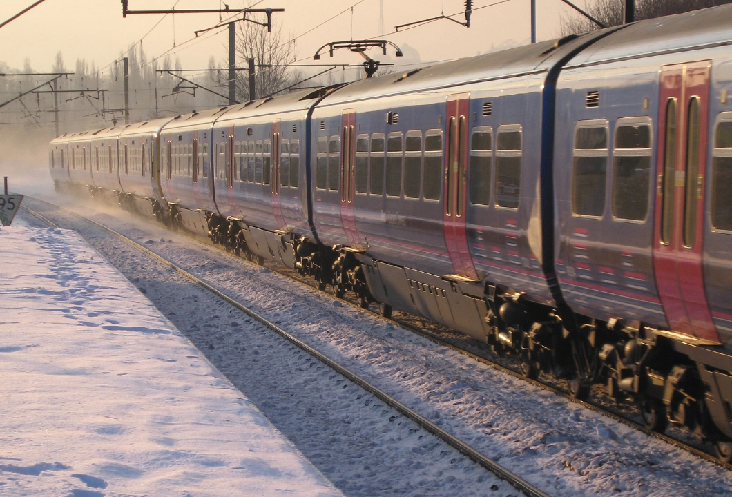 Photo of a blue and red train on a snowy track beside a snowy platform with an orange sky behind it.