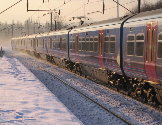 Photo of a blue and red train on a snowy track beside a snowy platform with an orange sky behind it.