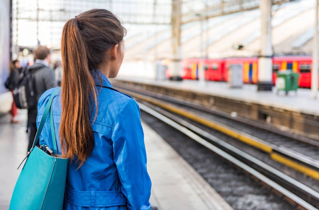 Photo of a train platform. The platform and people on the platform are blurred except for a woman in the foreground who stands with her back to the camera with long brown hair in a ponytail and a blue denim jacket. A red train is waiting at the platform opposite.