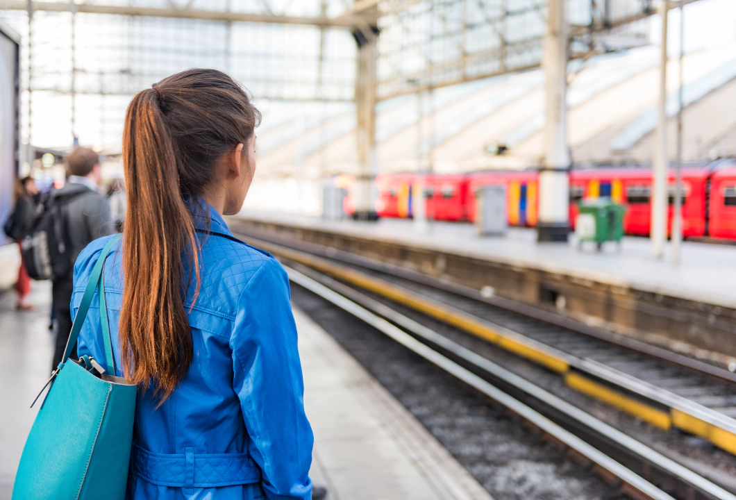 Photo of a train platform. The platform and people on the platform are blurred except for a woman in the foreground who stands with her back to the camera with long brown hair in a ponytail and a blue denim jacket. A red train is waiting at the platform opposite.