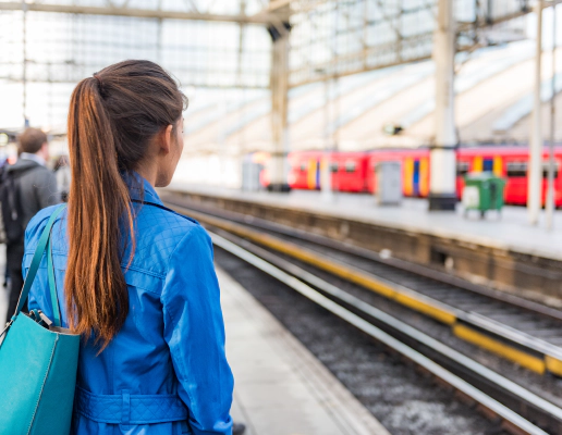 Photo of a train platform. The platform and people on the platform are blurred except for a woman in the foreground who stands with her back to the camera with long brown hair in a ponytail and a blue denim jacket. A red train is waiting at the platform opposite.