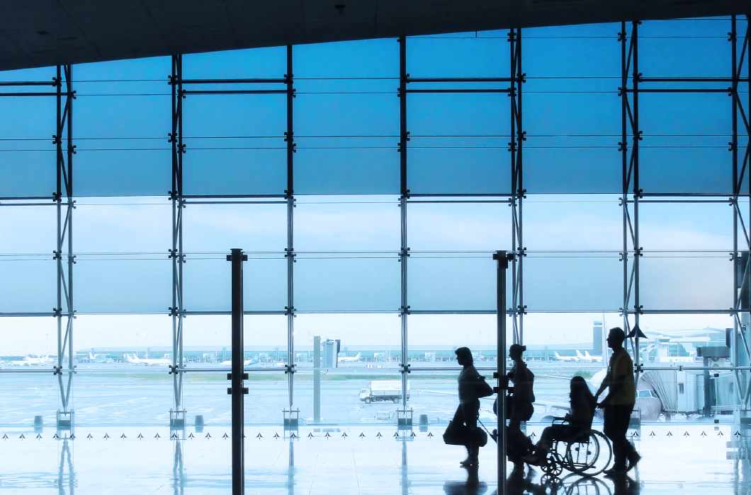 Four silhouettes of people in an airport setting, with a man and woman walking together with suitcases, followed by a wheelchair user being pushed by another person