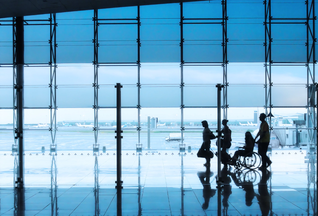 Four silhouettes of people in an airport setting, with a man and woman walking together with suitcases, followed by a wheelchair user being pushed by another person