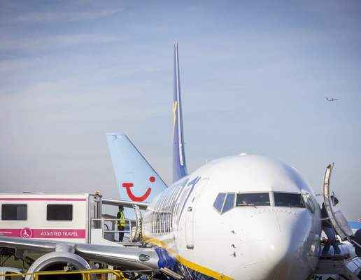 A zoomed in photograph of a Ryainair Boeing 747 plane from the front, with passengers disembarking by the front exit, by stairs, on the right, and an ambulift docking with the central exit of the plane on the left.