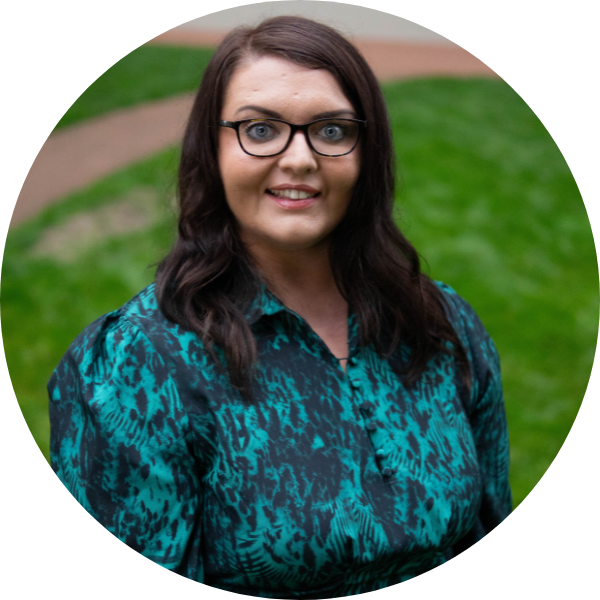 Circular photo of Emma Partlow, Transreport's Director of Consumer Engagement and Policy. Emma is a white woman with glasses and long dark hair, wearing a teal and black print dress, smiling in front of a green grass background.