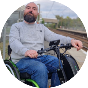 Joe, a white man with a brown beard and shaved head, sits in his wheelchair at a Railway Station.