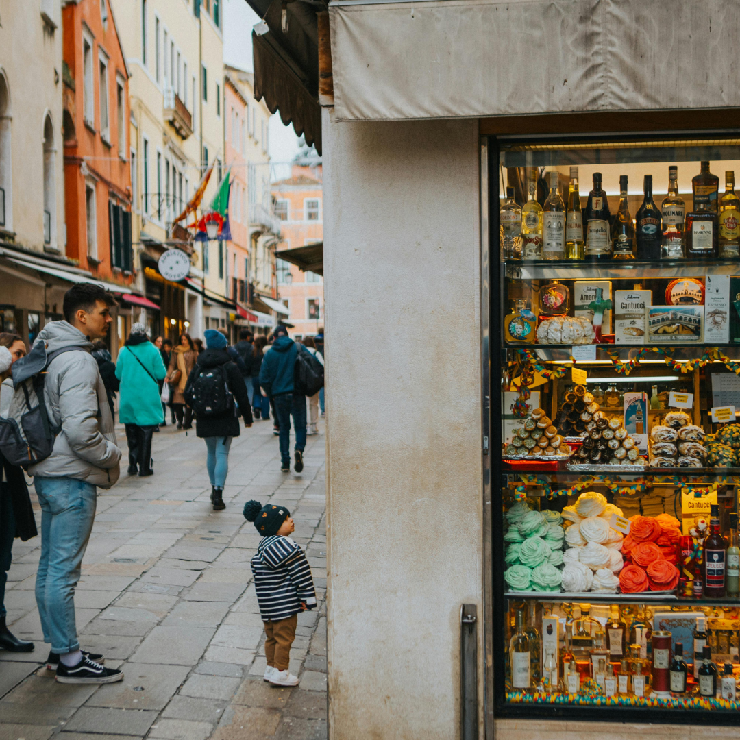 Photo of a family of three, a man, woman, and toddler, looking at a shop with a glowing windo display of deli food and wine products.