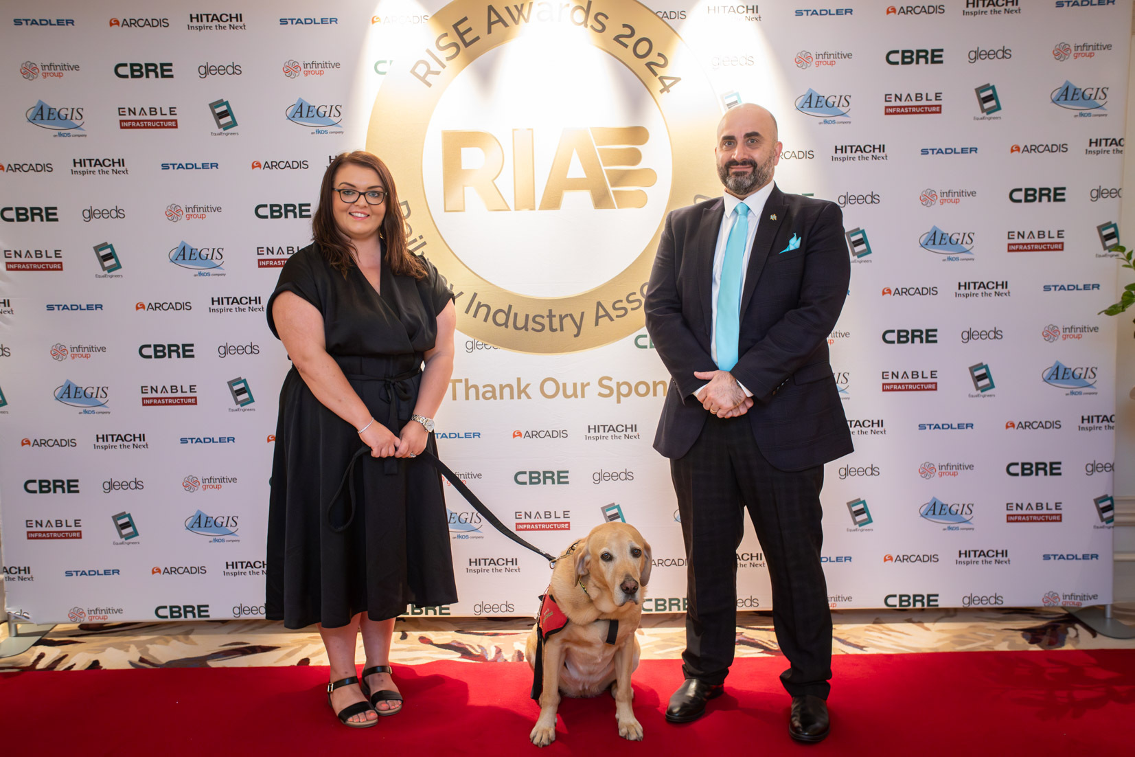 Emma Partlow, Transreport's Accessibility and Inclusion Manager, and Joseph Mastracci, Transreport's Commercial Manager, stand in front of the RIA Rise Awards 2024 sign on a red carpet. Emma is wearing a black dress and holding her assistance dog Luna on a black lead. Joseph is wearing a suit with a blue tie. They are both smiling.