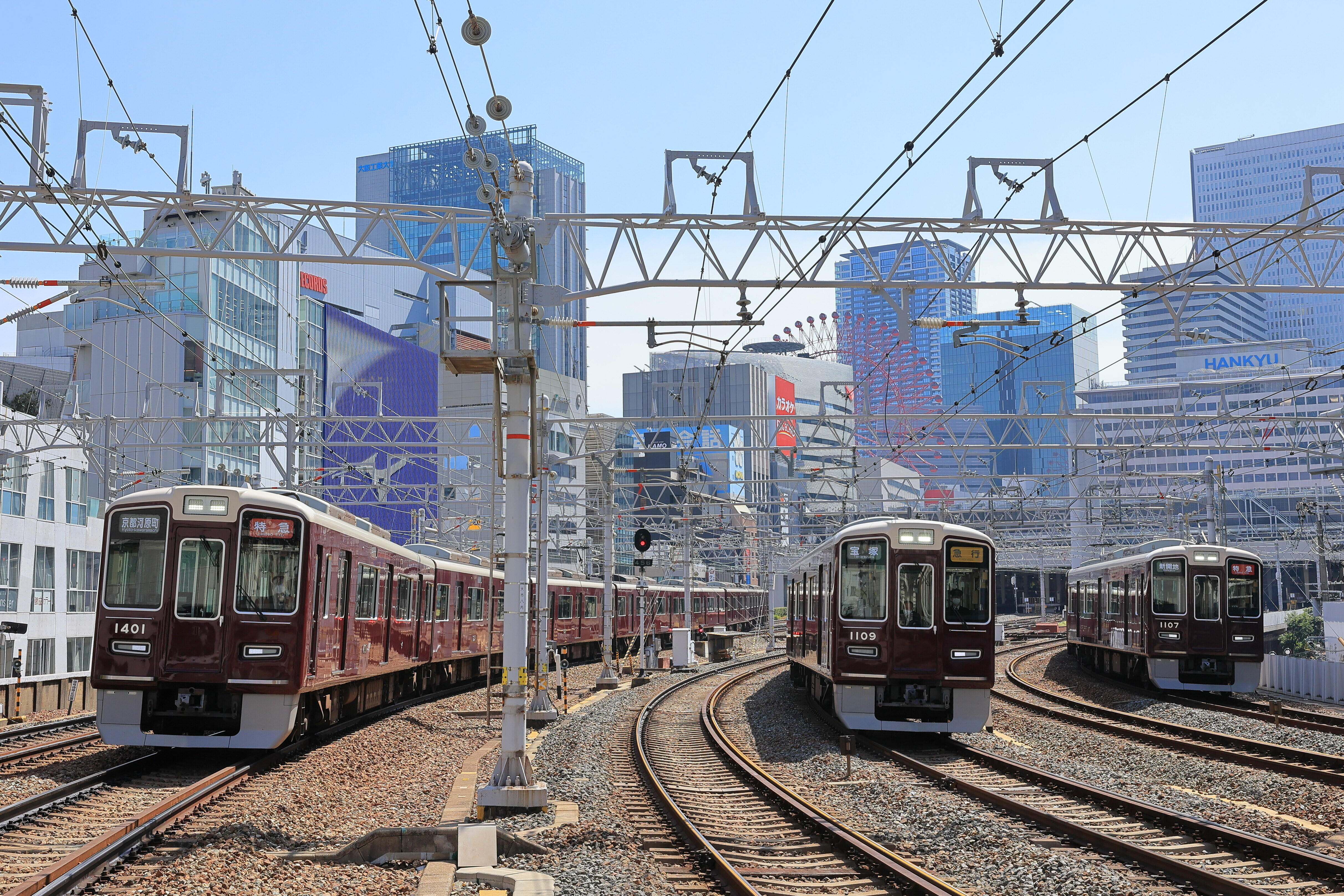 Image of Hankyu railway station. Three trains with multiple rail tracks surrounding them. There are buildings in the background.