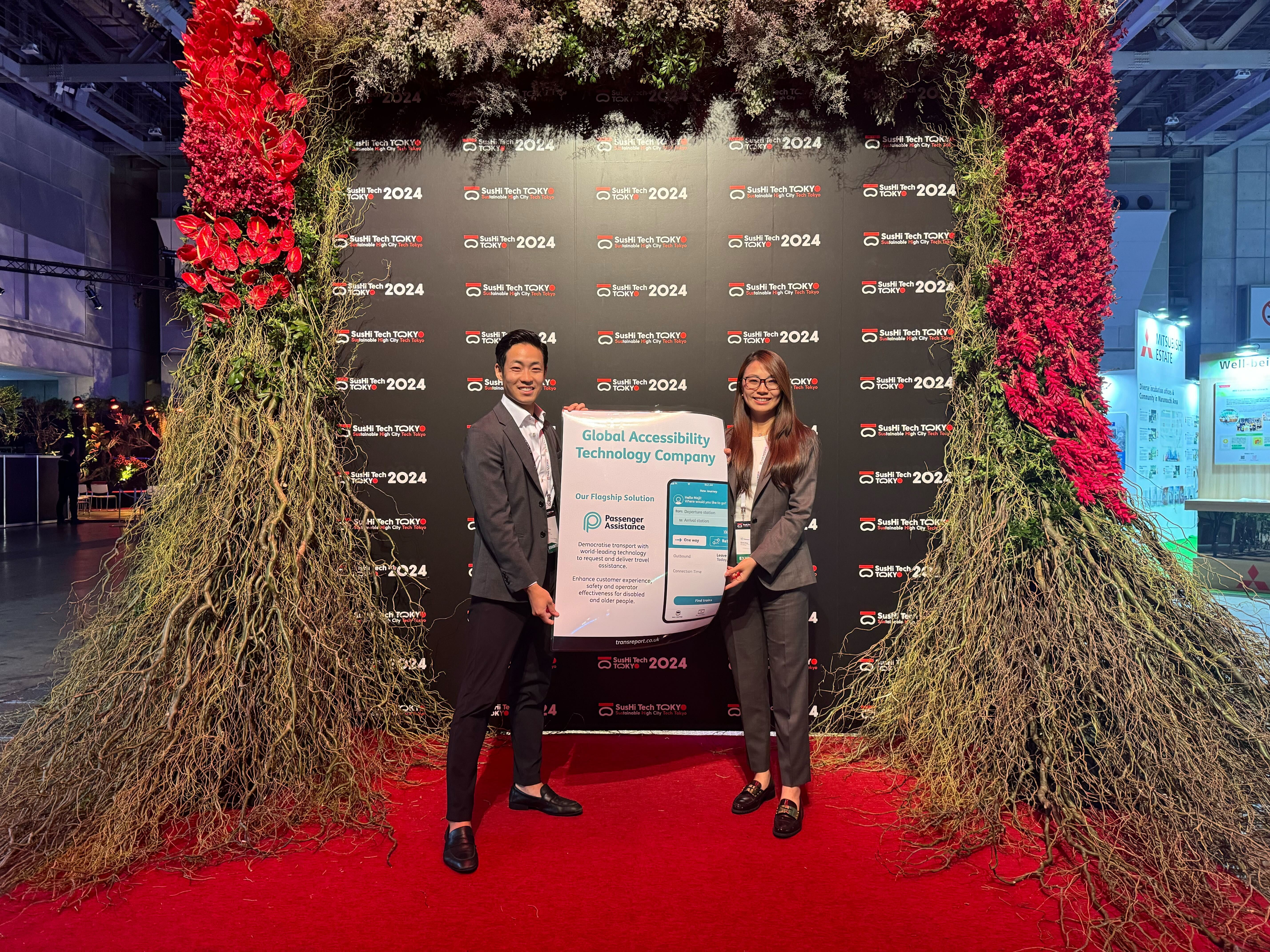 An East Asian man with dark hair, wearing a grey suit with a white shirt, holds a Passenger Assistance poster with an East Asian woman who has long brown hair, glasses and a grey suit. The poster has a teal heading on a white background Global Accessibility Technology Company. Beneath this is the teal text Our Flagship Solution above the Passenger Assistance logo. To the right on the poster is an image of a smartphone showing the Passenger Assistance app. The man and woman are smiling, standing on a red carpet in front of the SusHi Tech sign which is surrounded by vibrant flowers.