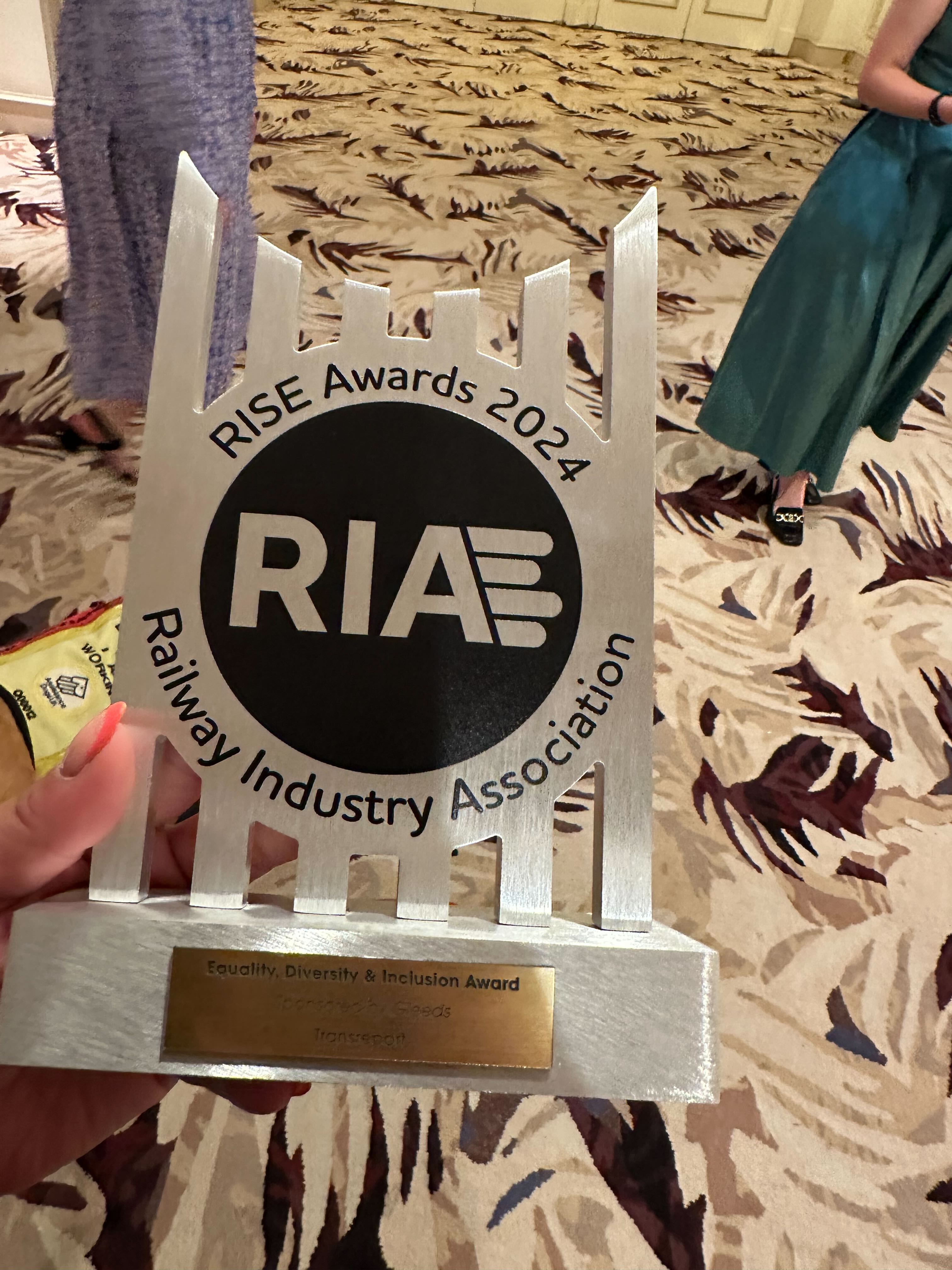 Transreport's award held over a patterned carpet. The award is a silver trophy with a black circle containing RIA in silver. RISE Awards 2024 curves around the top of the circle in black font. Railway Industry Association curves around the bottom. On the base of the award is a gold plaque on which is written Equality, Diversity & Inclusion Award.