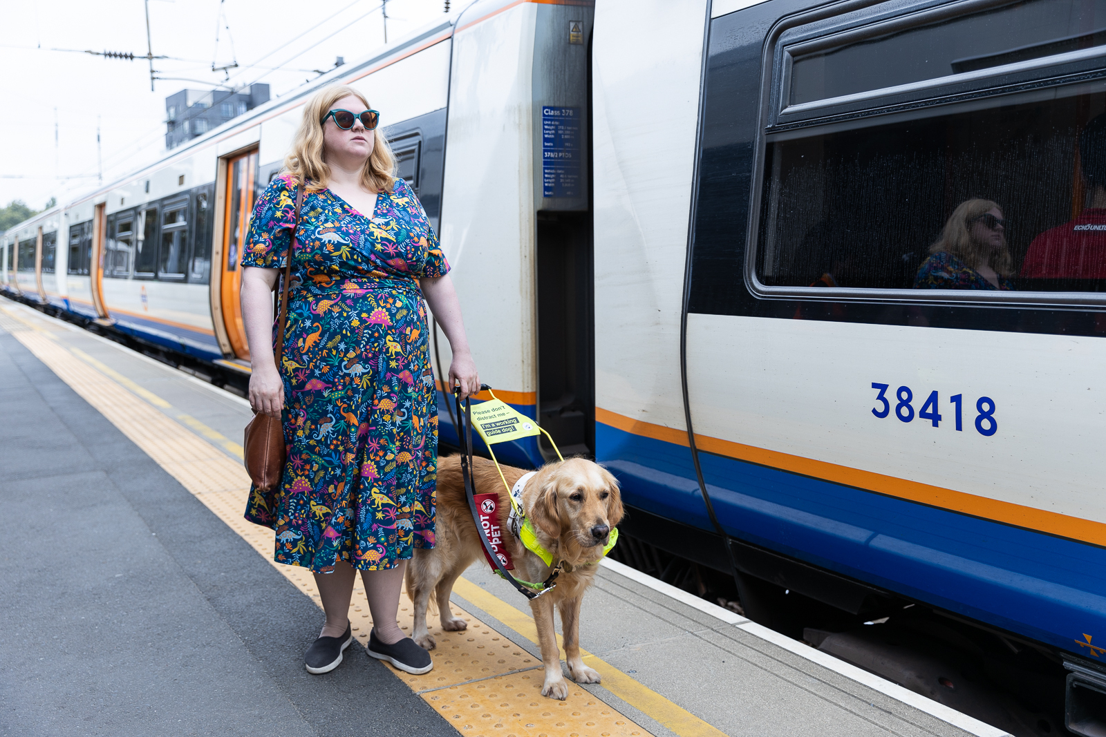 Amy with her guide dog Ava, standing on the platform beside a train. Amy has blonde hair and is wearing a dinosaur print dress.