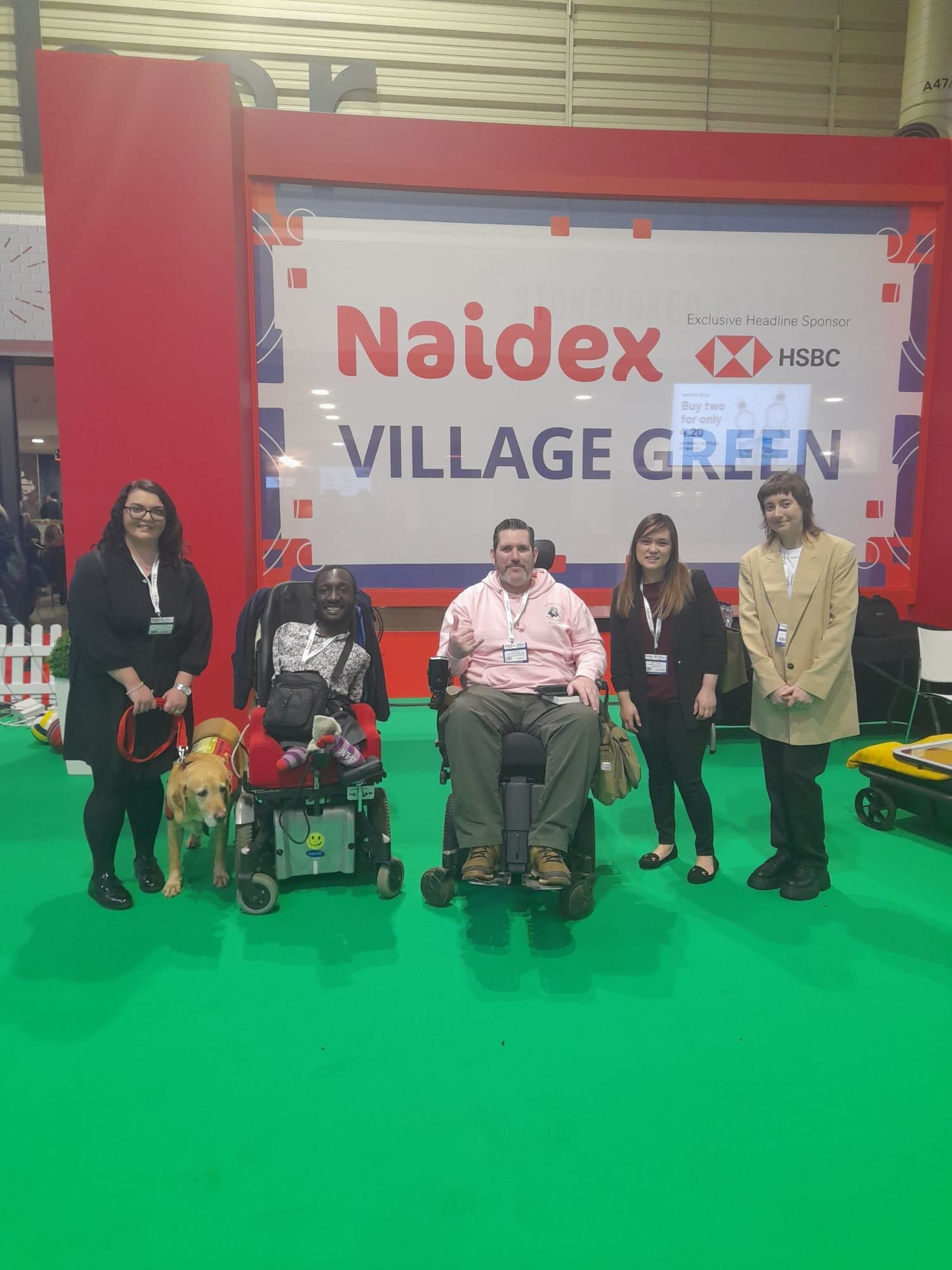 A photo of the Transreport team with Nick Wilson and Isaac Harvey, in front of the Naidex Village Green sign. From left to right - Emma Partlow, Transreport's Accessibility and Inclusion Manager. Isaac Harvey, MBE, President of Wheels and Wheelchairs. Nick Wilson, also known as the Disabled Adventurer. Linda Truong, Marketing Manager at Transreport. Mary Senier, Social Media and Communications Executive at Transreport.