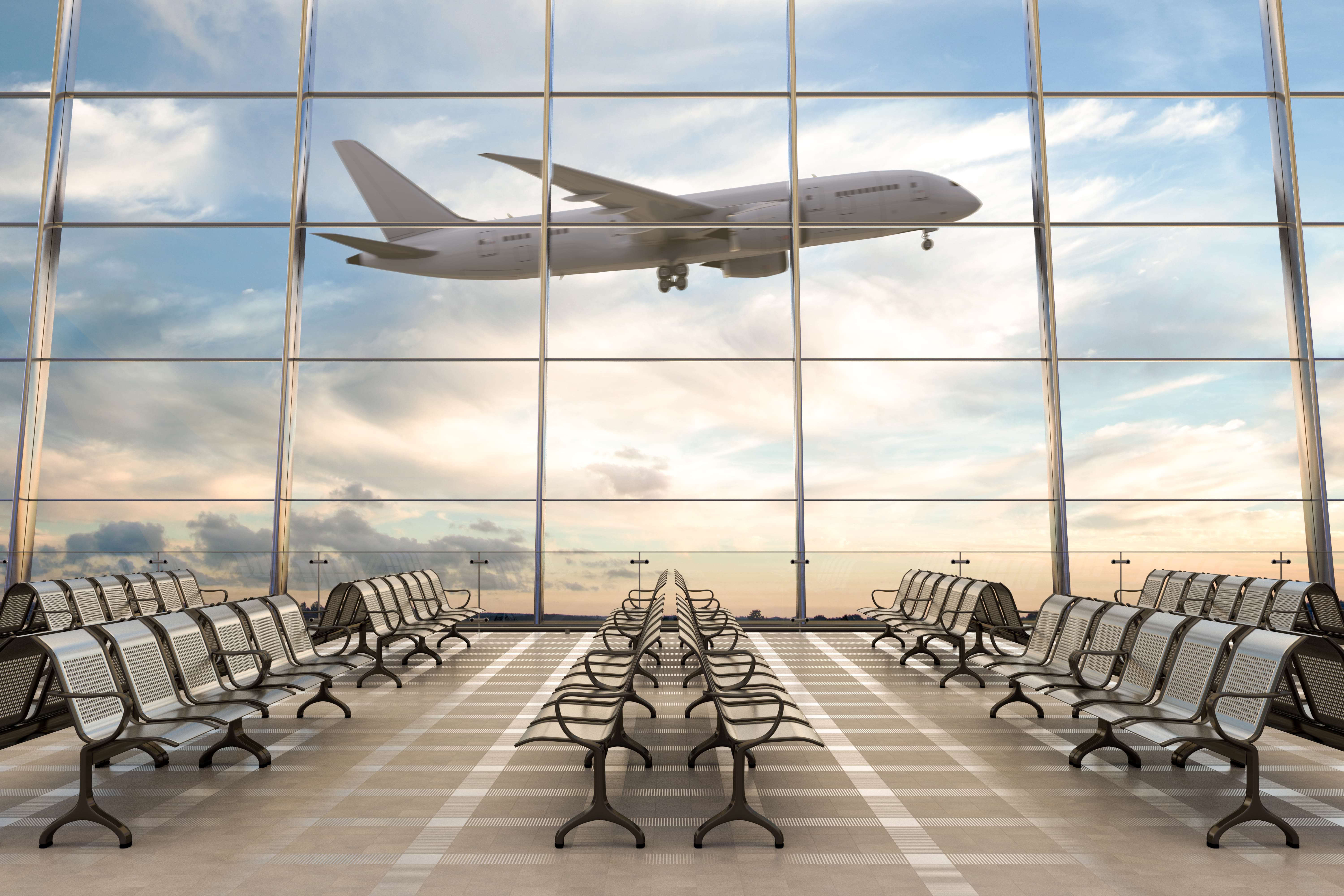 Photo of a white plane taking off beyond airport windows, against a blue sky with white clouds and gold sunrise. In the foreground of the photo is an empty airport lounge.