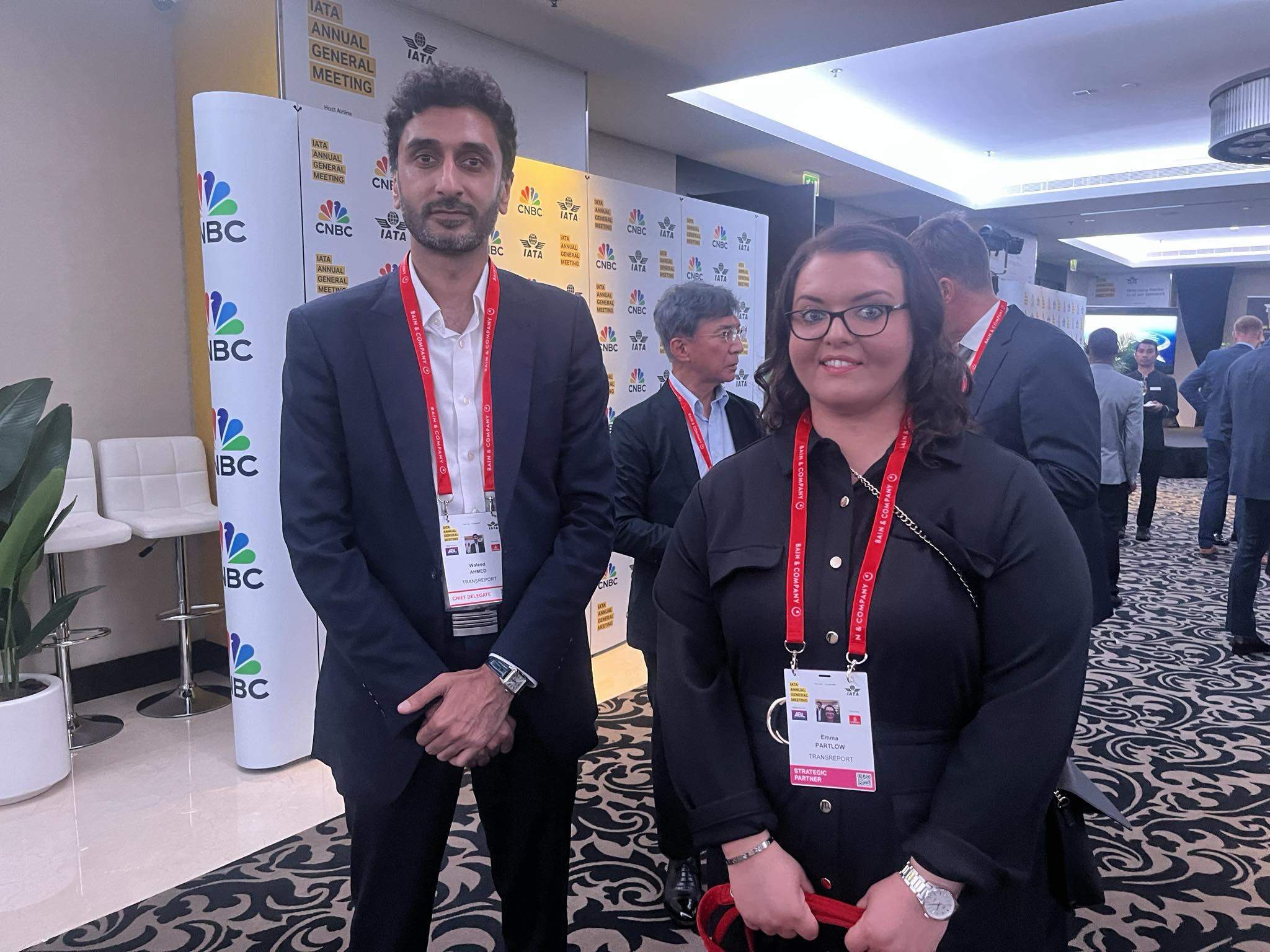 A photo of Waleed Ahmed, Transreport's COO and Emma Partlow, Transreport's Accessibility and Inclusion Manager standing in front of the IATA and CNBC sign. They are both smiling and wearing red and white lanyards. Waleed has black wavy hair and a beard and is wearing a navy suit with a white shirt. Emma has brown curly hair and glasses and is wearing a black dress.