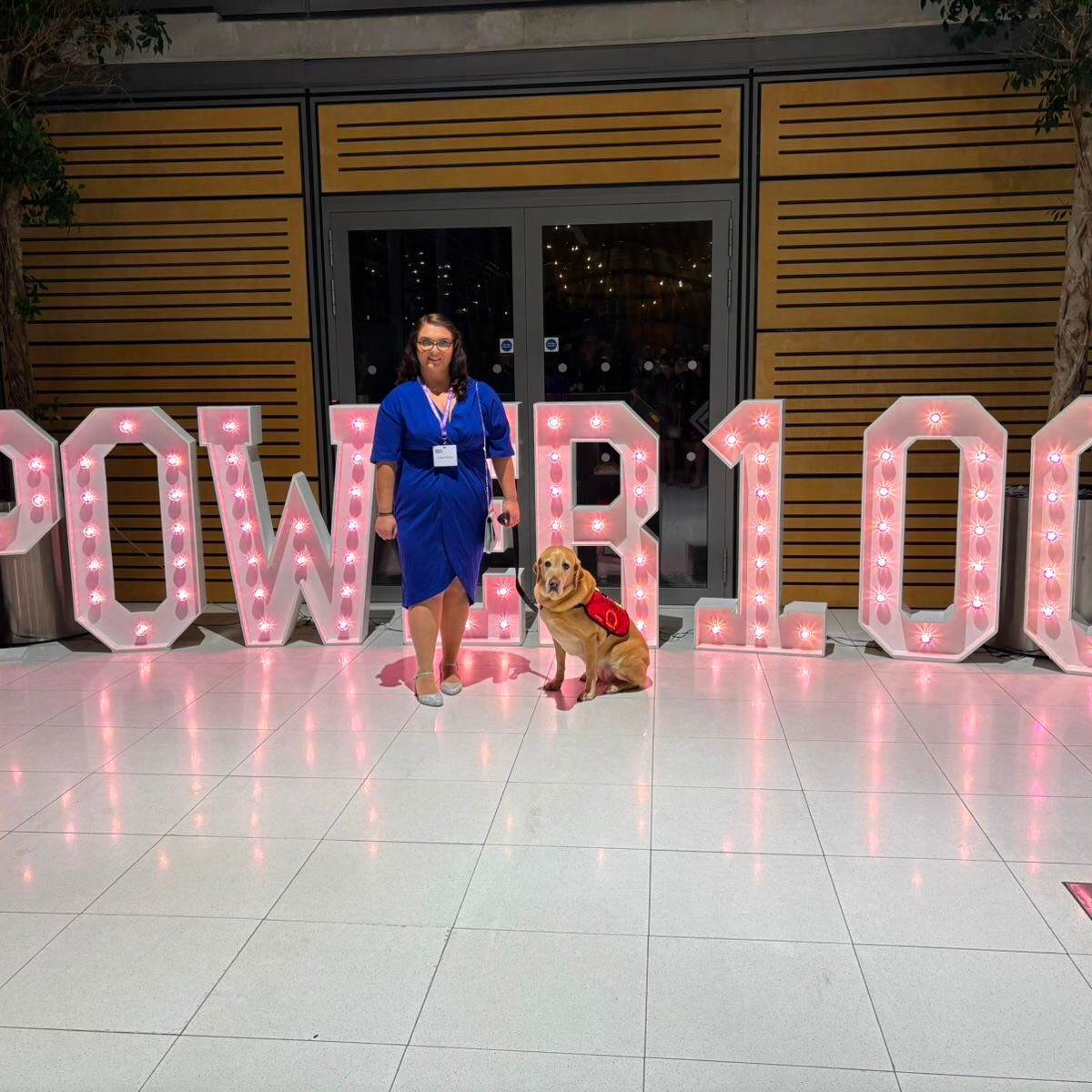 Photo of Emma Partlow, a white woman with long brown hair, smiling in a blue dress with sparkly silver heels, holds her assistance dog Luna, a golden labrador retriever with a red harness, on a black lead. Emma is standing on a white floor in front of the Power 100 sign, large solid white letters with pink LED lights.