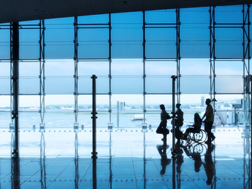 Four silhouettes of people in an airport setting, with a man and woman walking together with suitcases, followed by a wheelchair user being pushed by another person