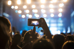 A photo of fans at a concert with one person taking a photo of the illuminated stage on their smart phone.