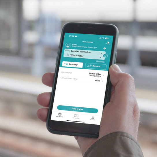 A photo of a person’s hand holding a smart phone showing the New Journey page in the PA App beside a train platform.